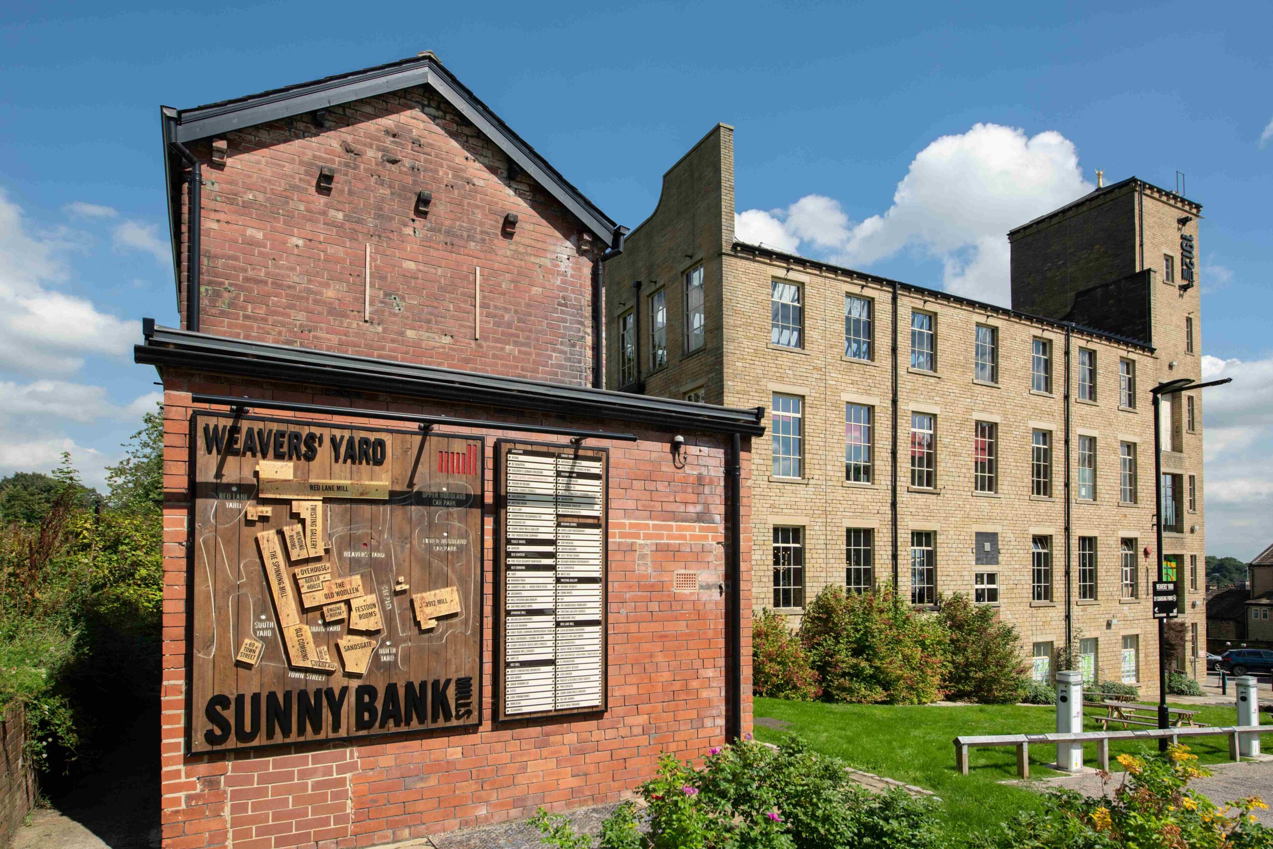 Photograph of Sunny Bank Mills from the outside. A sign on the wall reads 'Weavers' Yard Sunny Bank Mills' accompanying a wooden map of the site.