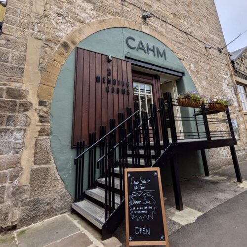 Metal stairs lead up to the 'Mending Rooms'. A sign over the door reads 'CAHM' and there is an A-frame sign board outside with a handwritten notice reading 'Candle Studio + Gift Shop. Summer Sale, up to 50% off end of line products. OPEN'