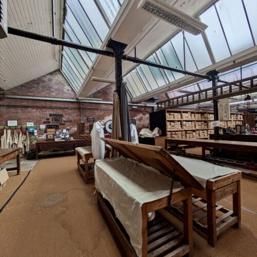 View into a long industrial room with skylights. There are long wooden benches, some covered with white cloth, and wooden shelves stacked full of carboard boxes.