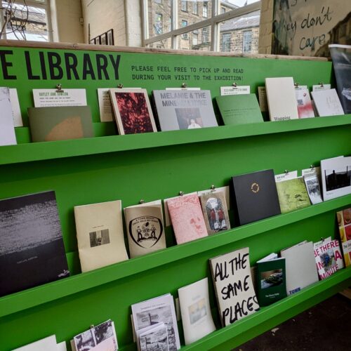 A bright green magazine rack with bold black lettering reading 'ZINE LIBRARY' is full of colourful paper zines.