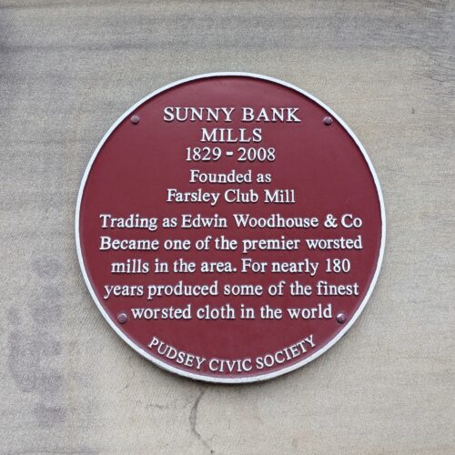 A red circular plaque hangs on a grey wall. On the plaque in white letters is written 'SUNNY BANK MILLS, 1829 - 2008. Founded as Farsley Club Mill. Trading as Edwin Woodhouse & Co. Became one of the premier worsted mills in the area. For nearly 180 years produced some of the finest worsted cloth in the world. PUDSEY CIVIC SOCIETY.'