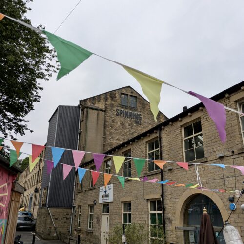 A view of part of the Sunny Bank Mill building from the outside. A sign on the outside reads 'SPINNING MILL'. Colourful bunting is strung across the street outside.