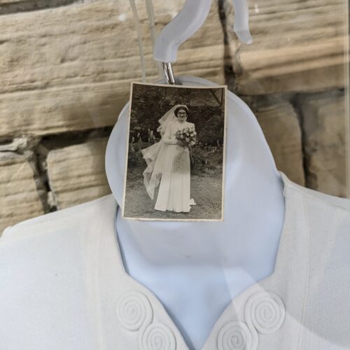 Close up of the neck of a hanger. There is a white garment on the hanger, and a black and white photograph attached to the hanger's hook of a woman in a wedding dress holding flowers.