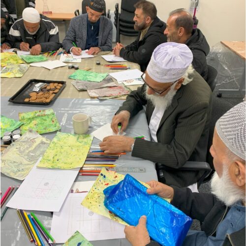 A group of older men, many wearing kufi caps, sit around a table, working with marbled paper.