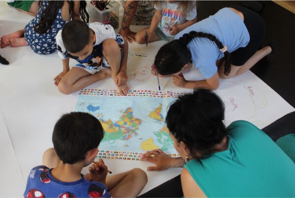 Young children and an adult crouch on the floor around a large map. Under the map, the floor is covered in paper for them to draw on.