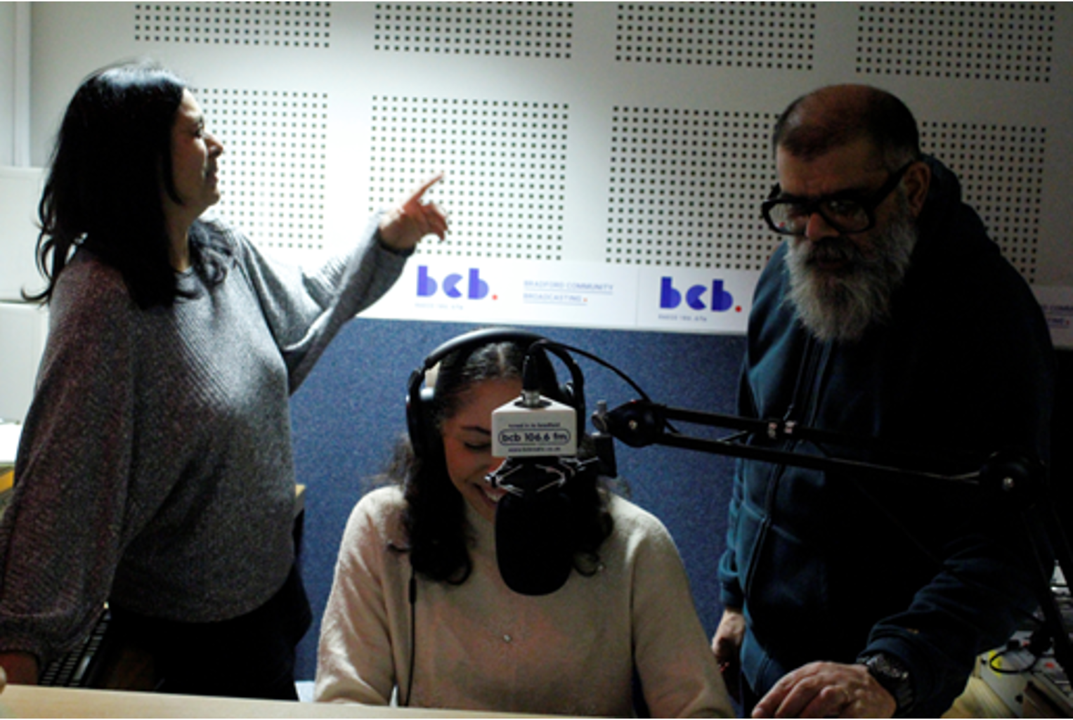 Three people in a radio studio, one wearing headphones in front of a microphone. The logo in the background reads 'bcb'.