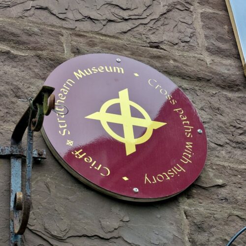 A close up photograph of a circular maroon and yellow sign bearing the Crieff and Strathearn Museum logo and the words 'Crieff and Strathearn Museum. Cross paths with history'.