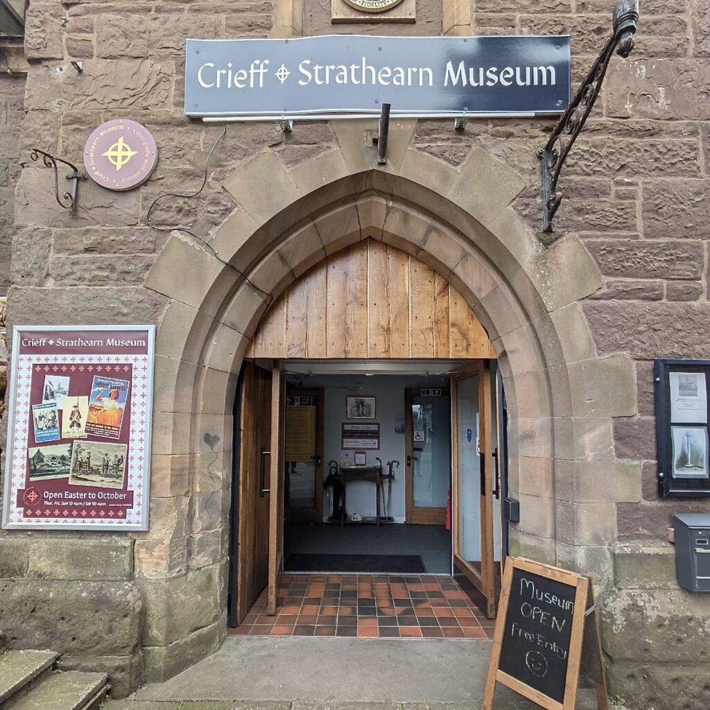 Photograph of the stone arched entrance to Crieff and Strathearn Museum. There is a sign bearing the name of the museum above the door, and boards on either side of the door promoting exhibitions.