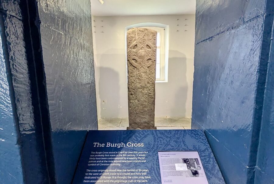 Photograph looking through a narrow viewpoint into the room where the Burgh Cross of Crieff is standing. There is an interpretation board in the foreground.
